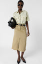 A woman wearing sunglasses, a light short-sleeve button-up shirt, a belted tan midi skirt with a front slit, black pointed shoes, and holding a black handbag stands against a plain background.
