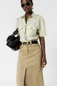 A person wearing a light short-sleeve button-up shirt, khaki skirt with front slit, black belt, gold jewelry, sunglasses, and holding a black handbag against a plain background.