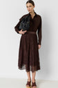 Woman wearing a dark brown button-up shirt, lace midi skirt, and brown heeled sandals, holding a black handbag under her arm, standing against a plain white background.