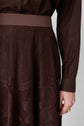 A person wearing a dark brown long-sleeve shirt and a matching brown lace skirt with floral patterns, shown from the waist down. Their hand with gold rings rests at their side.