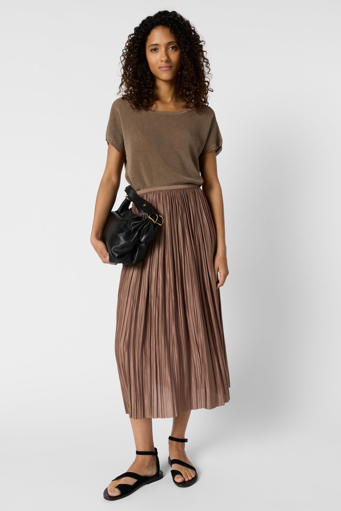 A woman with curly hair wears a brown short-sleeve top, a pleated midi skirt, black sandals, and carries a black bag over her shoulder, standing against a plain white background.