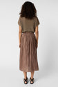 A person with curly hair stands facing away, wearing a short-sleeve brown top, a pleated brown skirt, and black sandals against a plain white background.