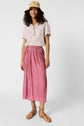 A woman wearing a tan sunhat, light pink short-sleeve blouse, pink pleated midi skirt, and tan sandals stands against a plain white background.