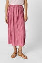 Person wearing a pink pleated midi skirt, a sleeveless white top with thin stripes, and tan strappy sandals, standing against a plain white background. Only the lower half of the body is visible.