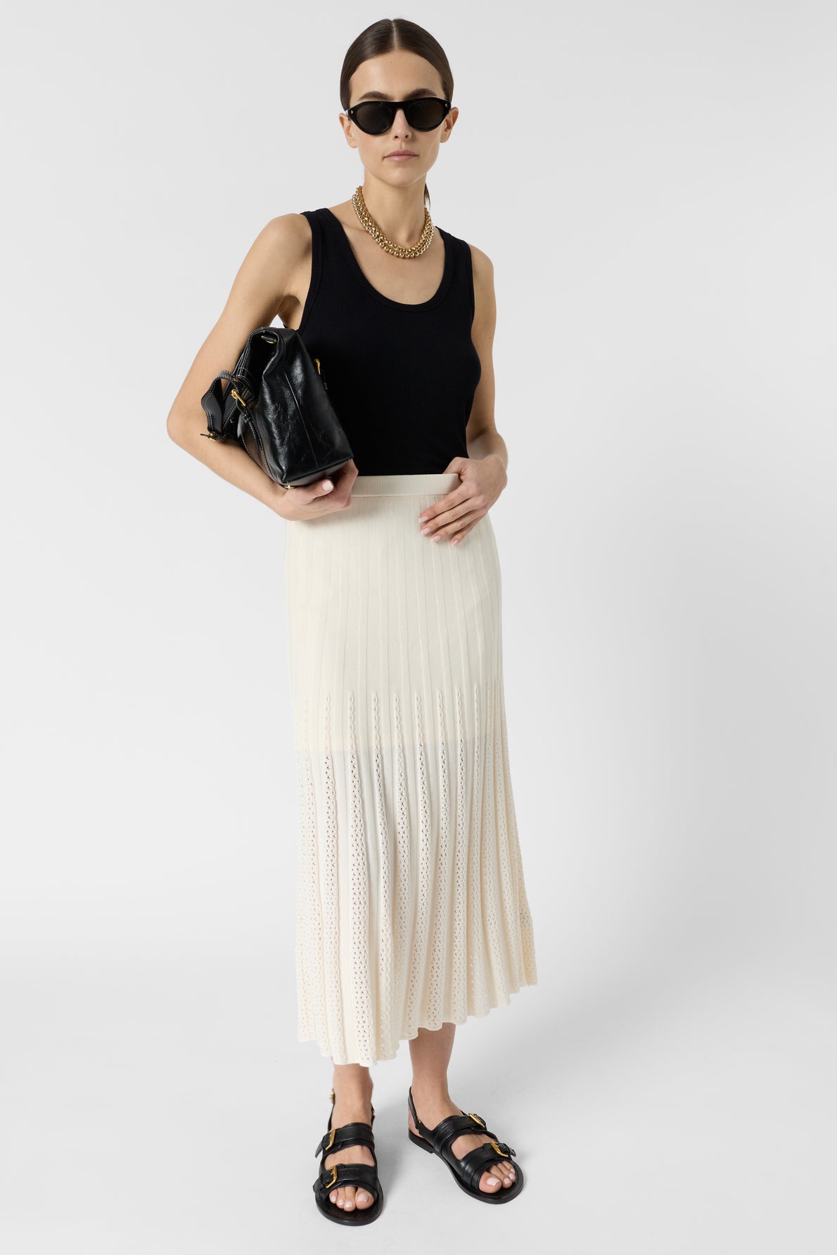 Woman wearing sunglasses, a black tank top, a gold chain necklace, a cream-colored jupe plissée, and black sandals, holding a black handbag, standing against a plain white background.