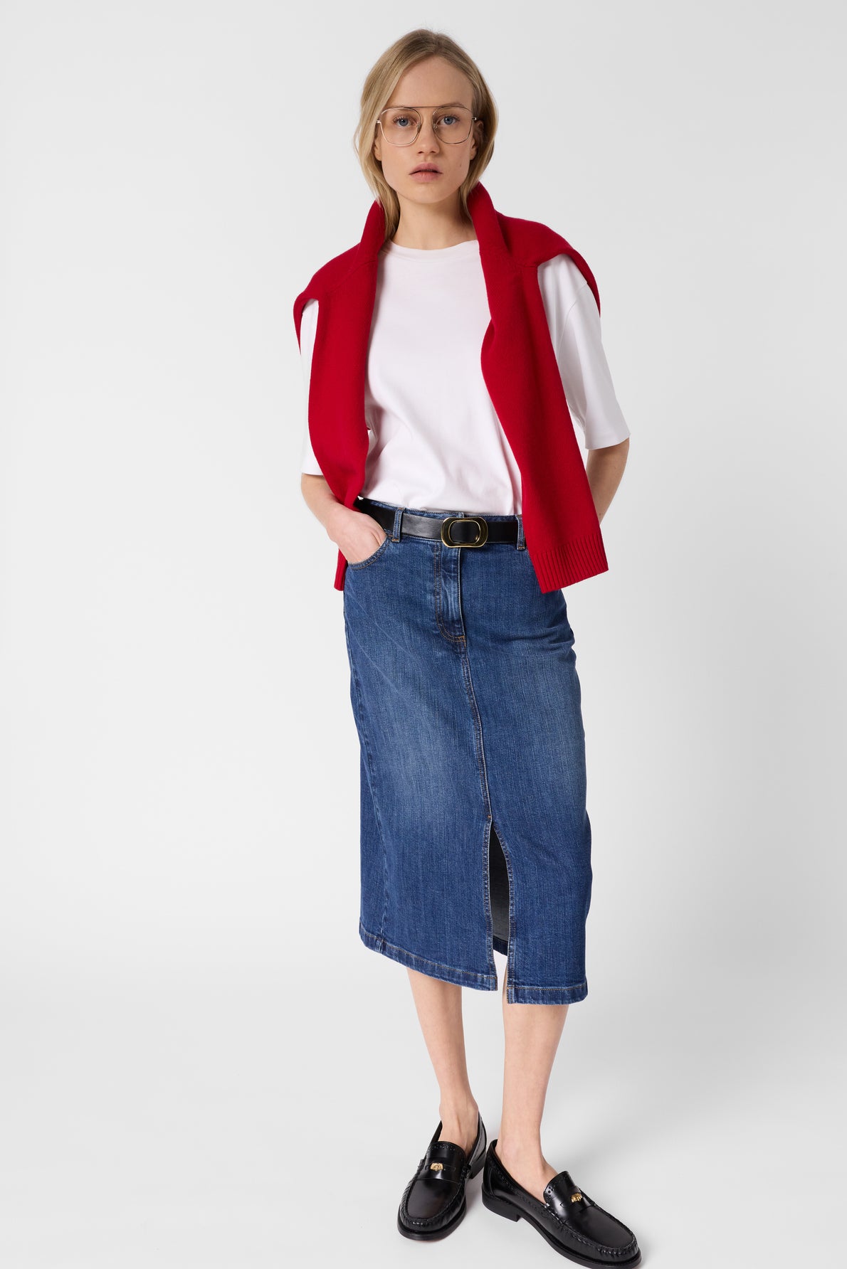 Person wearing glasses, a white t-shirt, a red sweater draped over their shoulders, a jupe droite en denim with a front slit, a black belt, and black loafers, standing against a plain white background.