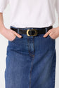 Person wearing a white shirt tucked into a jupe droite en denim, accessorized with a black belt featuring a large gold oval buckle. Hands are placed in the skirt’s pockets.