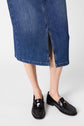 Person wearing a jupe midi femme in blue denim with a front slit, and black shiny loafers with metal details, standing on a white background.