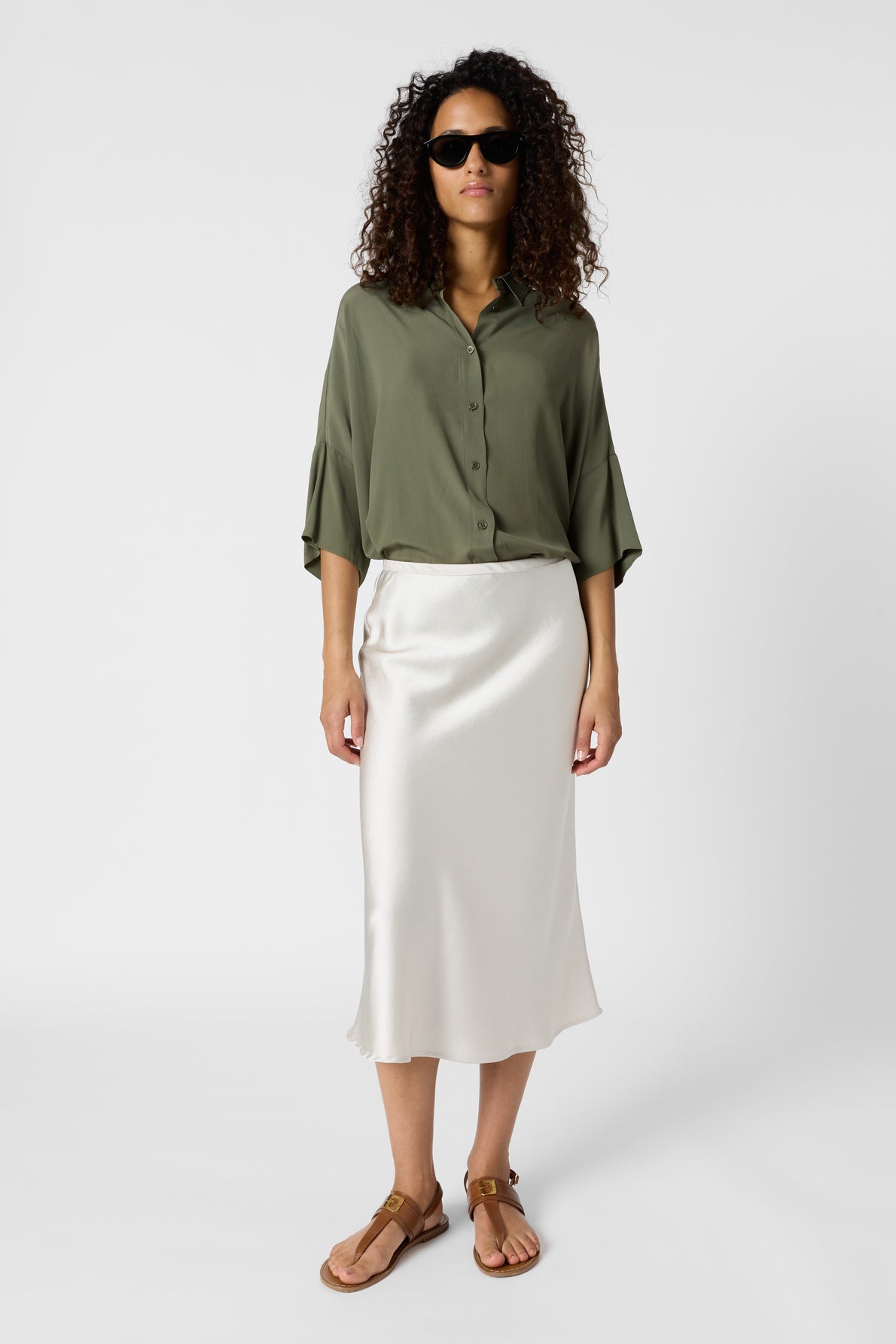 Woman wearing sunglasses, an olive green button-up shirt, a jupe satin écru midi skirt, and brown sandals stands against a plain white background.