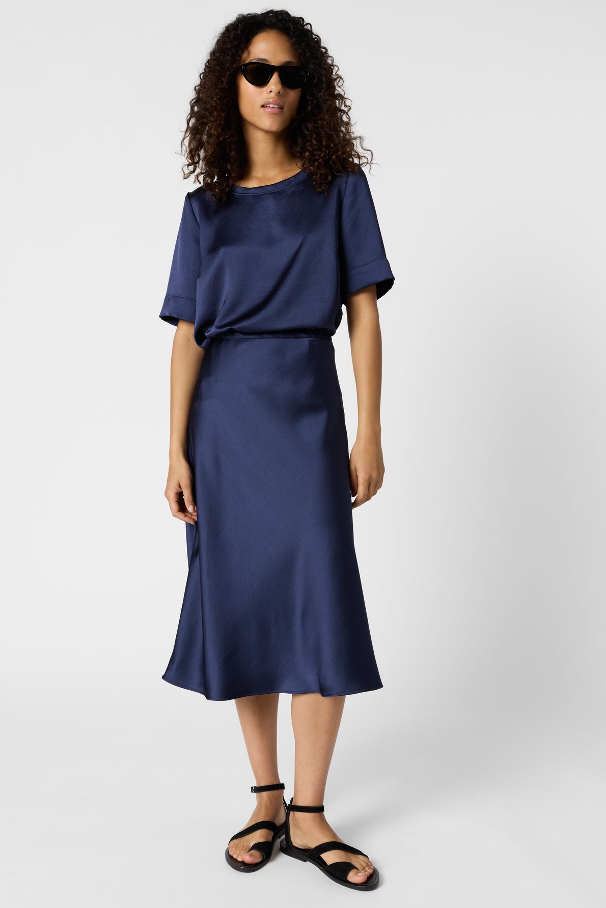 Woman with curly hair wearing black sunglasses, a short-sleeved navy blue satin top and matching skirt, and black sandals, standing against a plain white background.