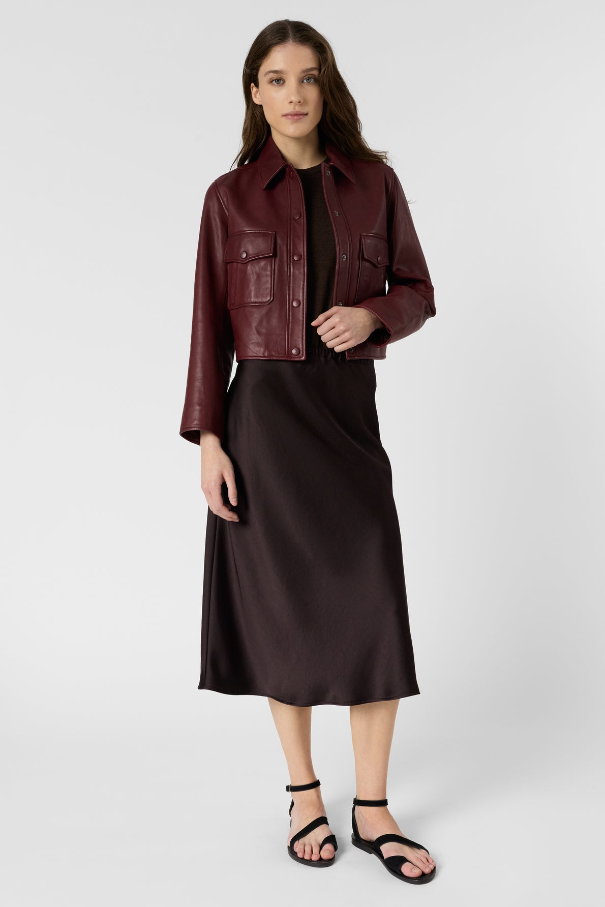 Woman wearing a burgundy leather jacket over a black midi dress and black sandals, standing against a plain white background.
