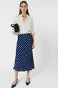 Woman standing against a white background wearing a cream blouse, a navy blue jupe midi motif à pois, black heels, and holding a black clutch bag.