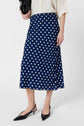 A person wearing a cream blouse and a navy blue jupe midi motif à pois, holding a black bag. They are also wearing black pointed-toe slingback heels.