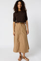 A woman wearing a dark brown short-sleeve top, a tan belted jupe camel midi skirt en coton, and brown sandals stands against a plain white background.
