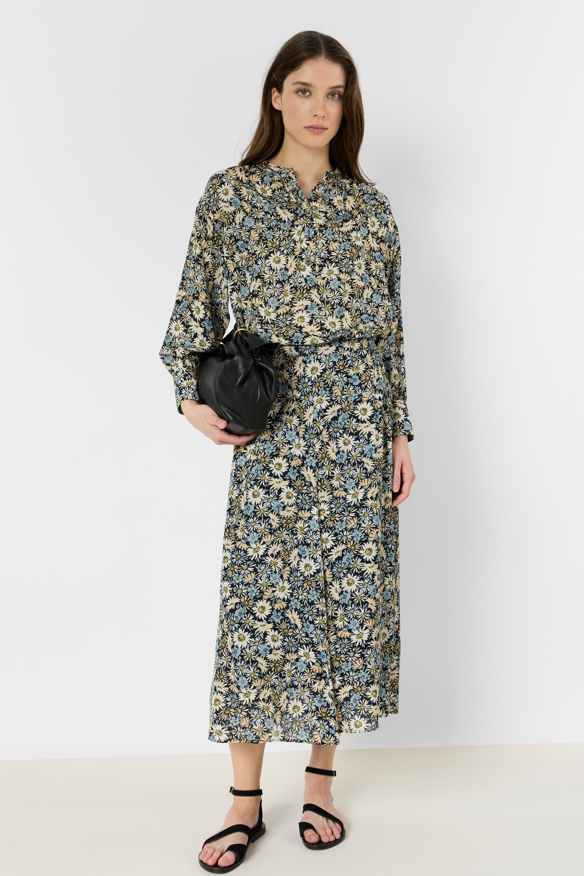 Woman standing against a white background, wearing a long-sleeved, floral-patterned dress with a jupe midi silhouette, black sandals, and holding a small black bag.