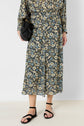 Person wearing a jupe midi motif floral, matching floral top, black sandals, and holding a small black purse. The jupe fluide features a blue, white, and yellow floral pattern on a dark background. The photo is cropped at the shoulders.