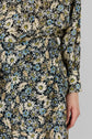 Close-up of a person wearing a black dress with a cream, yellow, and blue daisy motif floral print. The garment features long sleeves with button cuffs and a button-down detail on the hip, creating the look of a jupe midi. The background is a plain, light grey wall.