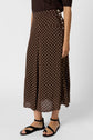 A person wearing a brown jupe à pois, ankle-length with white polka dots, paired with black ankle-strap sandals and a dark top. The skirt features buttons along the hip.
