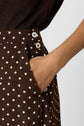 A person wearing a brown jupe midi à pois with white buttons has their right hand in the pocket. The fluid skirt is paired with a dark brown top.
