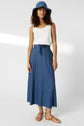 Woman in a white sleeveless top, blue high-waisted skirt with a belt, blue hat, and tan sandals stands facing the camera against a plain light background.
