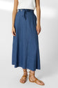 A person wearing a white sleeveless top, a high-waisted blue maxi skirt with a tie belt, and tan strappy sandals, standing against a plain background.