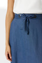 A person wearing a white top and a high-waisted blue skirt with a front tie and grommet details. Only the lower torso and one arm are visible.