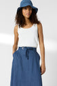 Woman wearing a blue sunhat, white sleeveless top, and a blue high-waisted skirt with a belt, standing with one hand in her pocket against a plain background.