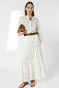Woman in a long white eyelet dress with a brown braided belt, holding a tan purse, and wearing brown sandals, standing against a plain white background.