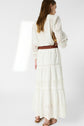 Woman standing with her back to the camera, wearing a long white eyelet dress, brown braided belt, and brown sandals, holding a small brown bag against a plain white background.