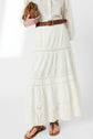 Woman wearing a long white eyelet skirt with a brown braided belt, holding a brown bag. She is also wearing a white eyelet blouse and brown sandals. Only her body from shoulders down is visible.