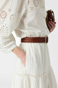 A person wearing a white eyelet dress with a braided brown belt and holding a small brown purse. One hand is in the dress pocket. Only the torso and part of the arm are visible.