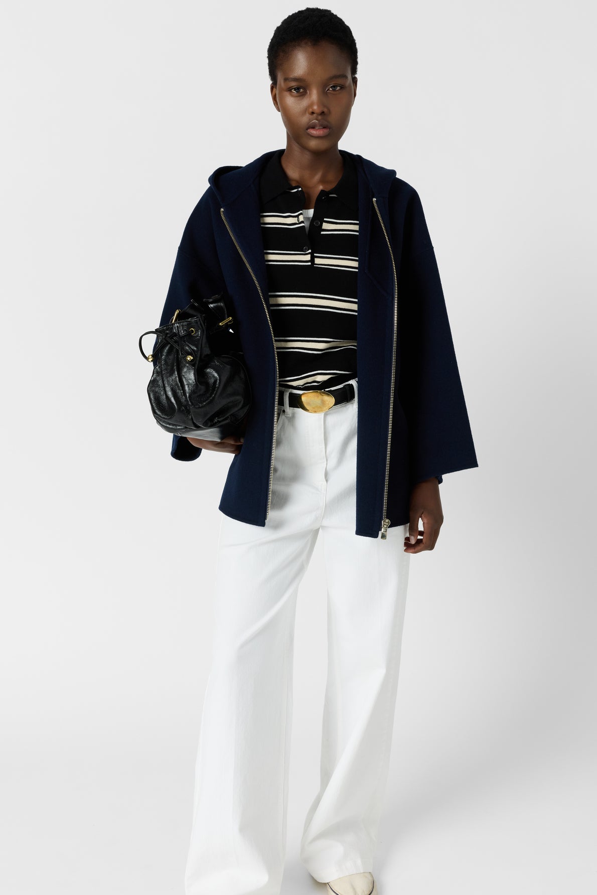 A person stands against a plain background, wearing a navy manteau laine femme jacket over a black-and-white striped polo, white pants, and holding a black handbag. They have a serious expression and short hair.