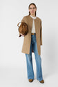 A woman stands wearing a tan coat, white sweater, blue flared jeans, and brown shoes. She holds a brown handbag and looks directly at the camera against a plain white background.