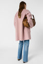 Woman standing with her back to the camera, wearing a pink coat, blue jeans, and brown shoes, holding a large brown handbag under her left arm against a plain white background.