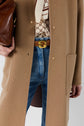 A person wearing a tan coat, patterned shirt, blue jeans with a brown belt featuring a gold buckle, and holding a brown handbag.