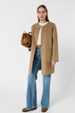 A woman in a tan coat, white top, blue flared jeans, and brown shoes stands holding a brown bag. The background is plain white.