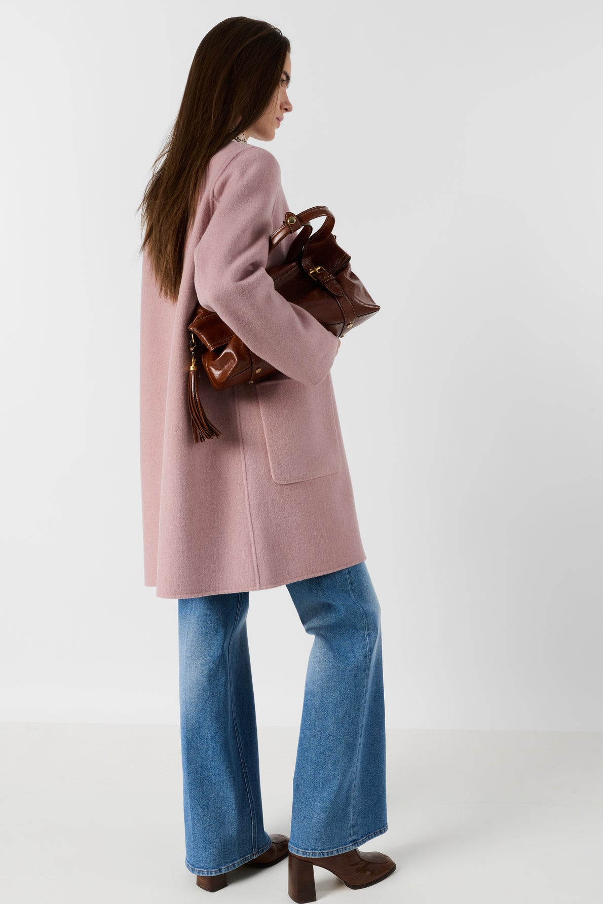 Woman with long brown hair, wearing a pink coat and blue wide-leg jeans, stands sideways holding a brown leather bag. She is in front of a plain white background.