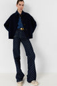 A person poses in a dark buttoned jacket, blue denim shirt, patterned high-waisted wide-leg jeans, white sneakers, and a black belt with a large gold buckle, standing against a plain white background.