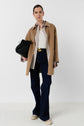 A person wearing a tan coat over a striped shirt, dark blue jeans, and white sneakers, holding a black handbag, stands against a plain white background.