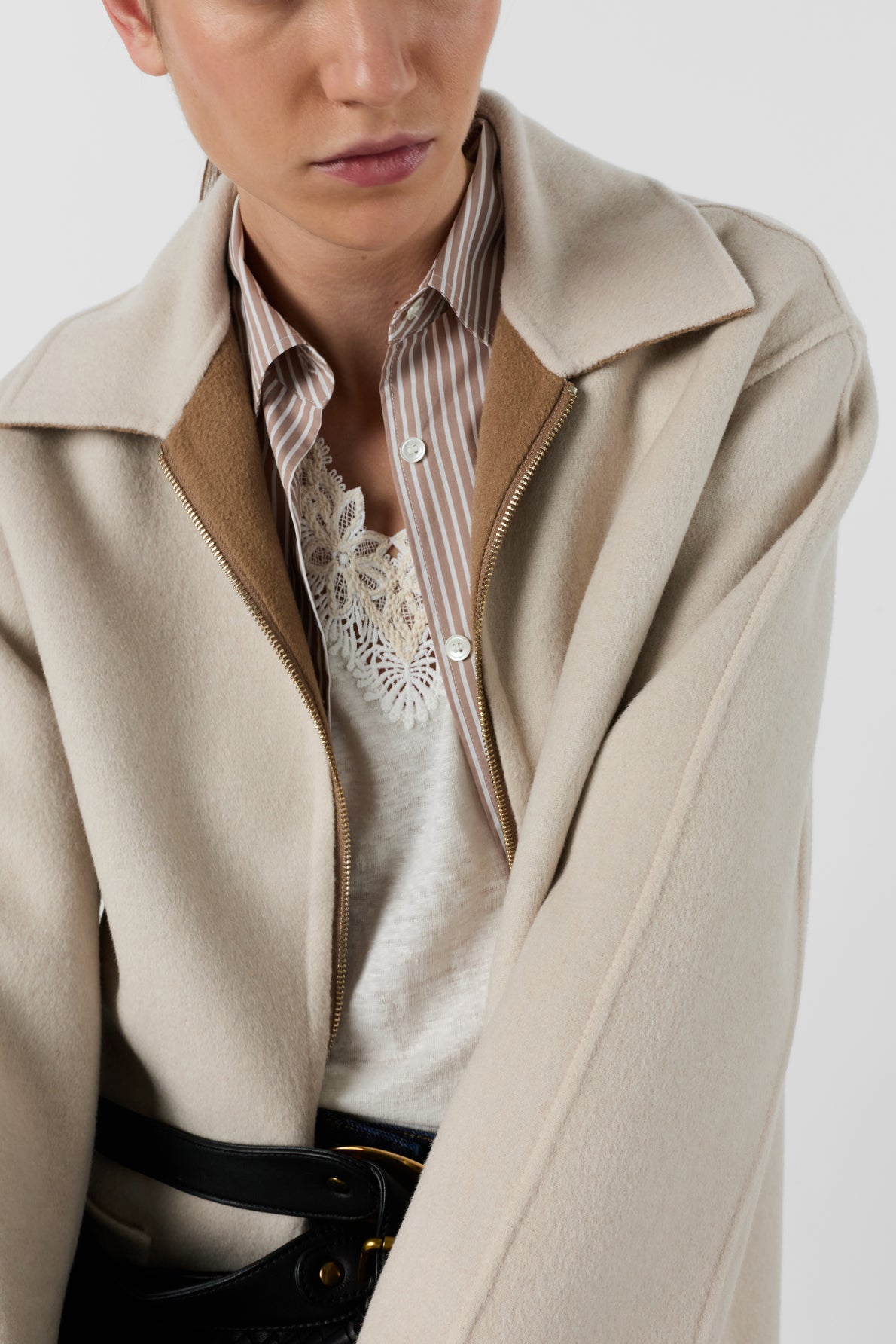 A person wearing a beige coat over a striped shirt and lace-trimmed white top, with the coat partially unzipped and black belt visible, against a plain background.