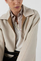 A person wearing a beige coat over a striped shirt and lace-trimmed white top, with the coat partially unzipped and black belt visible, against a plain background.