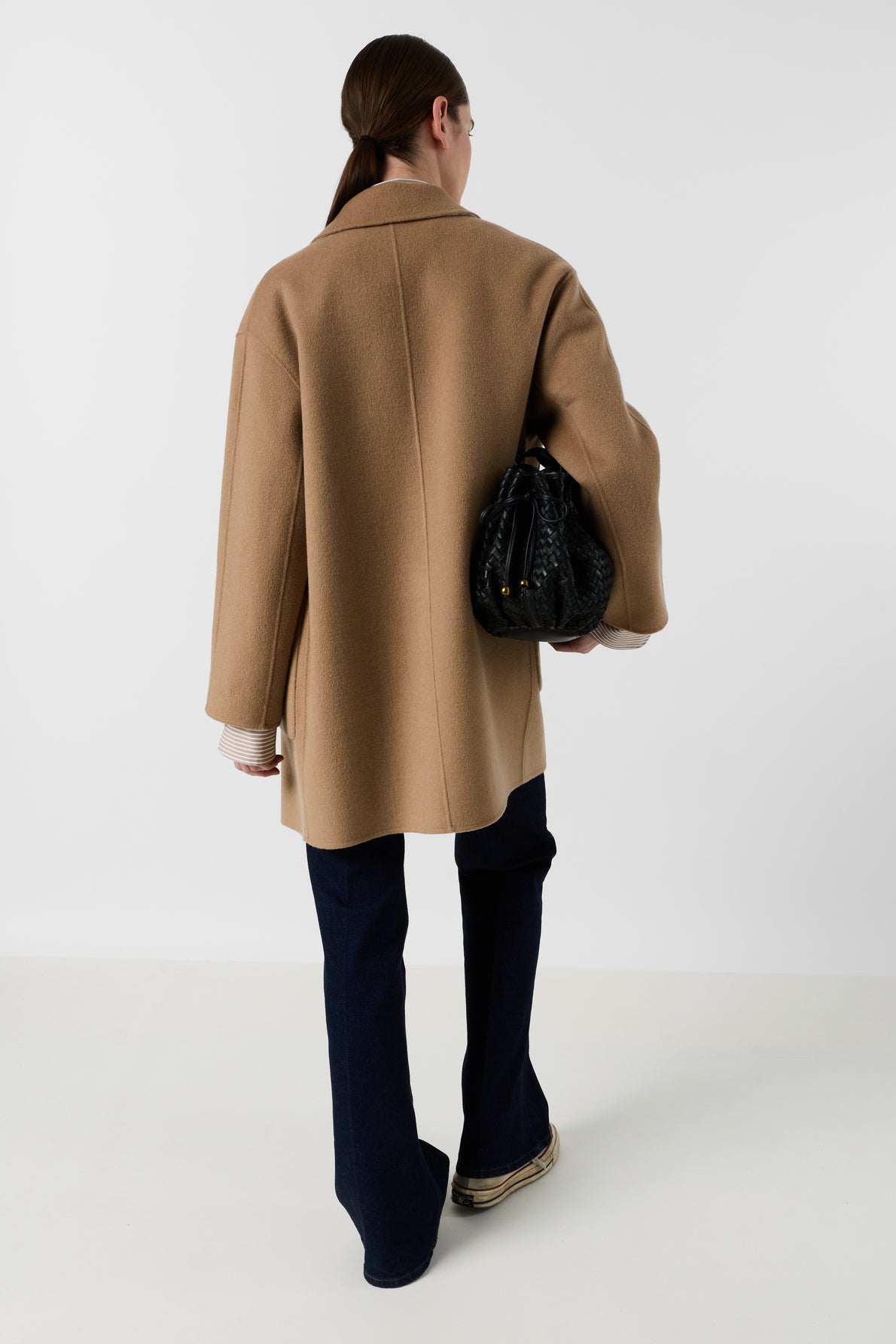 Person with long brown hair in a low ponytail, wearing a tan coat, dark pants, and white shoes, stands facing away while holding a black handbag on their left side against a plain white background.