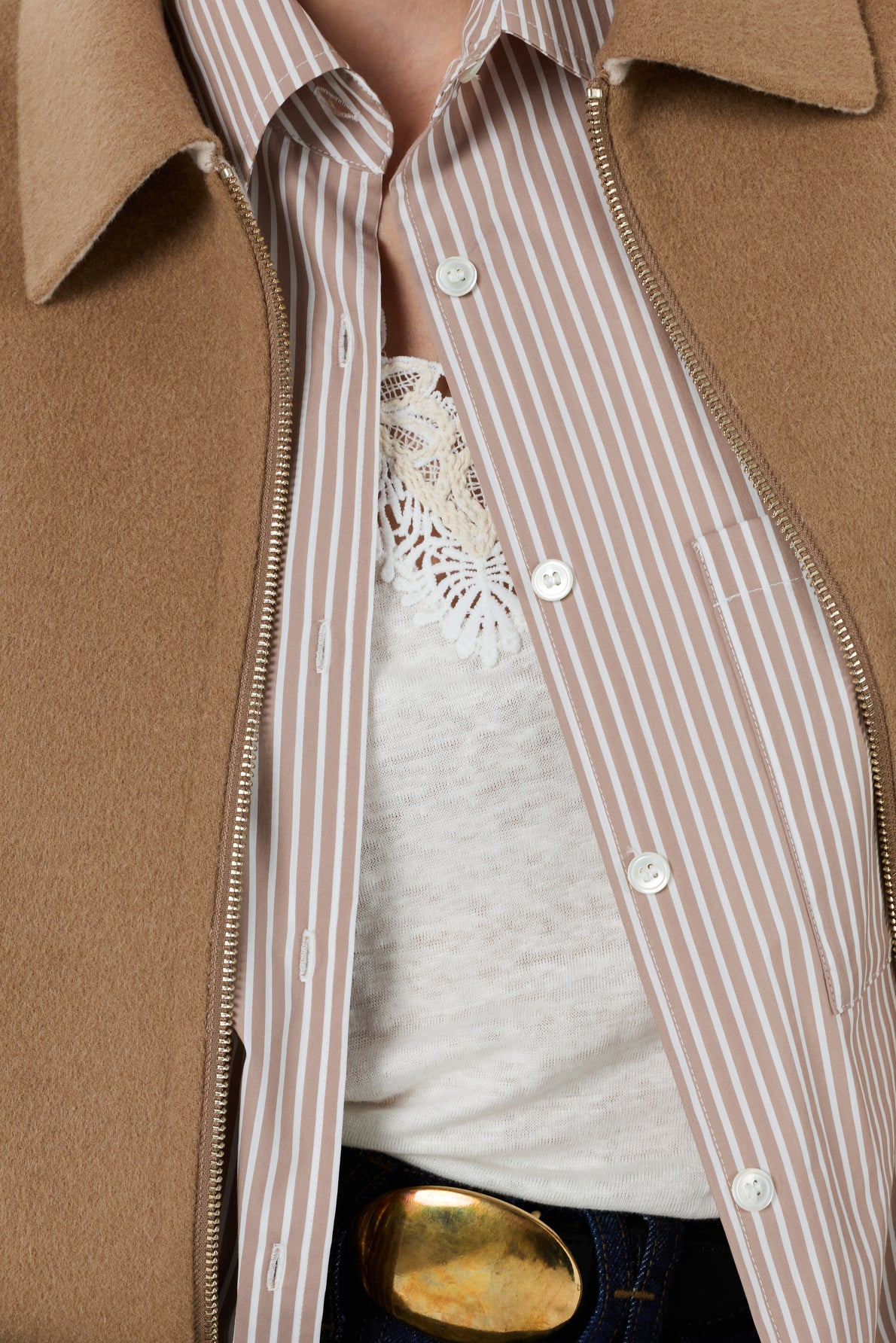 A close-up of a person wearing a tan coat, a brown and white striped button-up shirt, a white lace-trimmed top, and dark jeans with a gold belt buckle. Only the torso is visible.