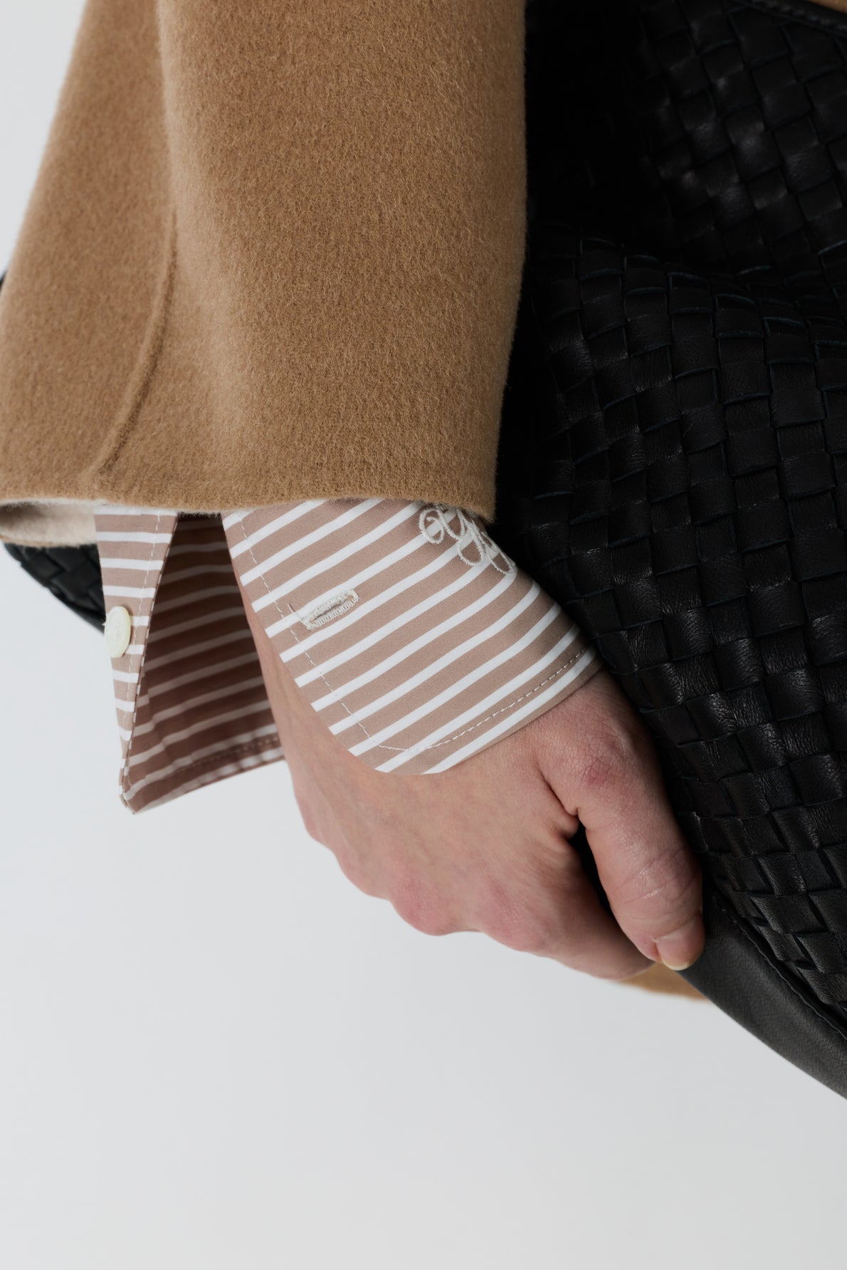 A person wearing a tan coat and a striped shirt cuff holds a textured black woven bag.