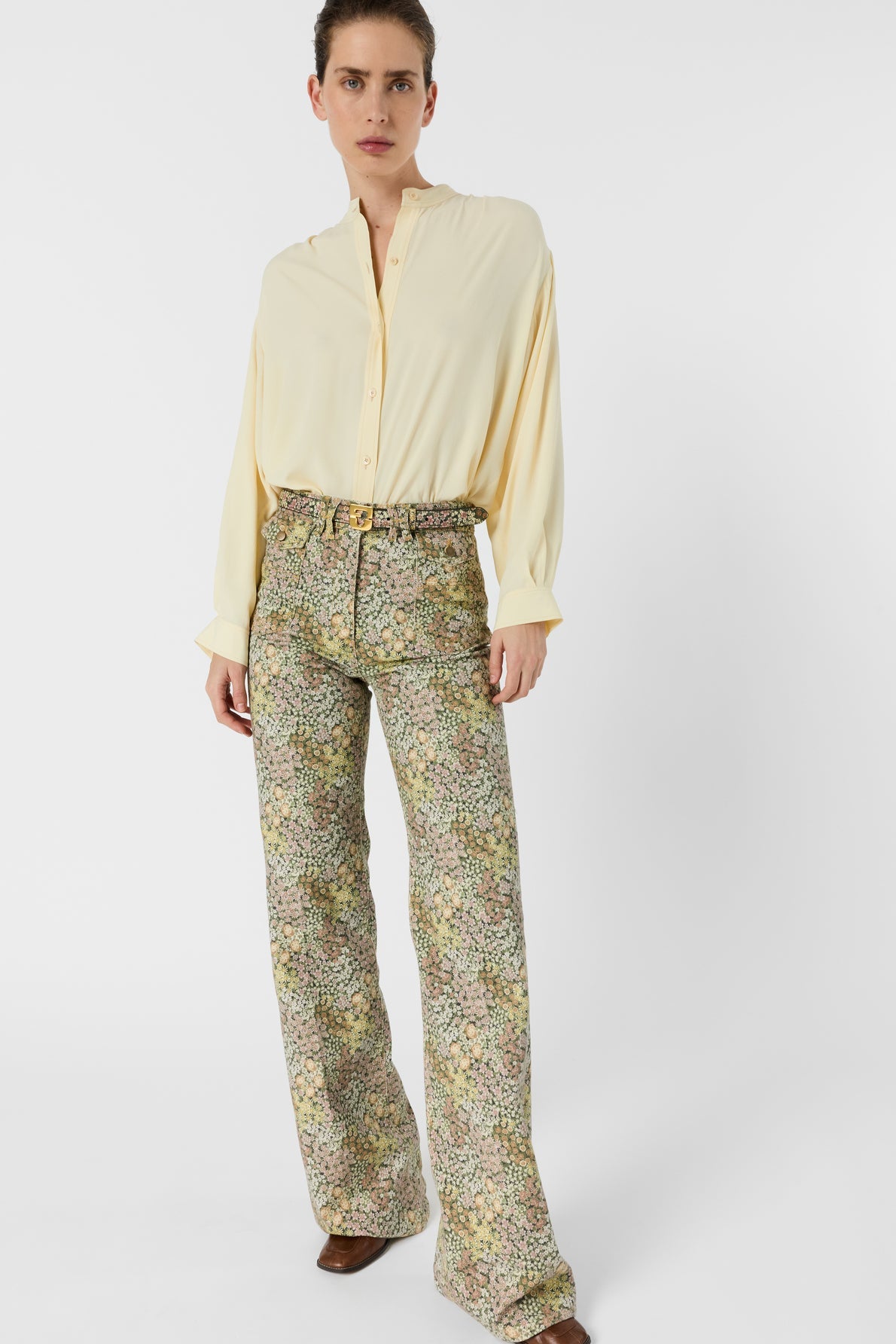 A person stands against a white background wearing a loose, light yellow button-up shirt and high-waisted, wide-leg motif floral pantalon flare with a belt and brown shoes.
