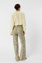 A person stands facing away, wearing a light yellow long-sleeve blouse and high-waisted pantalon flare jean ANNA with a green and yellow motif floral pattern, against a plain white background.
