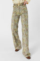 Person wearing high-waisted, wide-leg pantalon flare with a vibrant motif floral and a matching belt, paired with a light yellow shirt and brown pointed-toe shoes. The image is cropped above the shoulders, highlighting the stylish jean ANNA-inspired look.