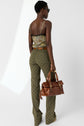 Person wearing a floral top, green patterned pantalon monogramme, and sunglasses stands holding a brown leather handbag. They are facing away from the camera against a plain white background.