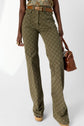 Woman wearing a high-waisted, olive green, patterned flared pantalon monogramme with a brown belt, holding a brown handbag. Only her torso and legs are visible. She also wears gold rings and platform shoes.