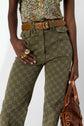 A person wearing green patterned pantalon monogramme, a brown belt, a floral top, gold rings, and holding a brown leather bag. The photo is cropped from the chest down.
